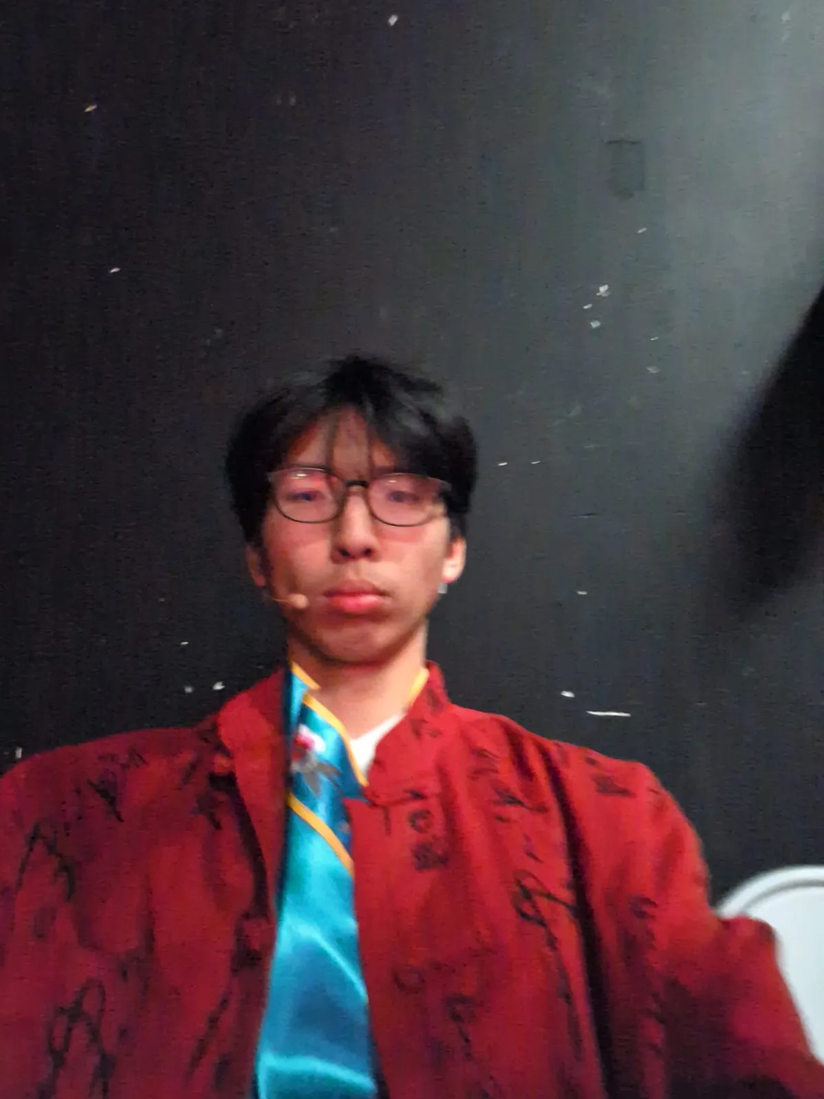 Ian in a red robe and teal tie in costume, wearing a stage mic