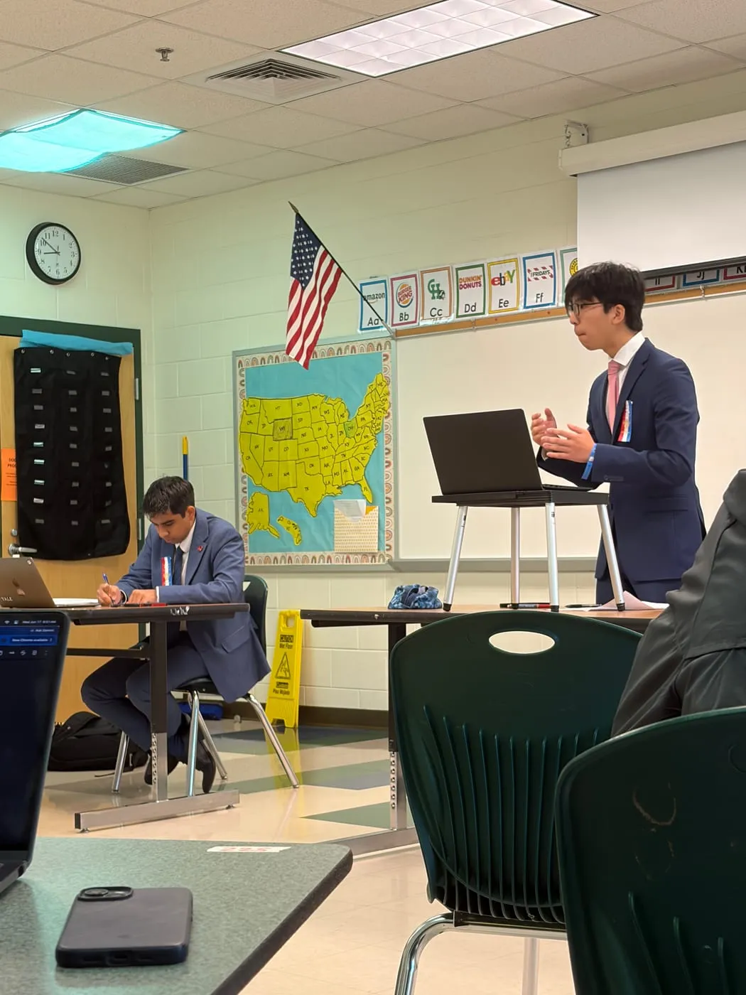 Ian speaking from a laptop stand in a debate round, with a judge seated in front of him